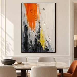 Vertical abstract artwork in orange, black, white, charcoal, and yellow, displayed in a dining room in a black frame.