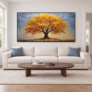 A broad golden autumn tree beneath a gray-blue sky in a black frame, displayed above a sofa in a living room.