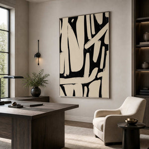 Silver framed monochrome abstract artwork behind a desk and cream reading chair beside bookshelves.