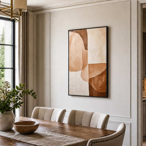 Black-framed earth-tone abstract painting above cream dining chairs beside a large window and wooden table.