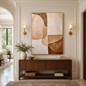 Gold-framed cream and caramel painting between wall sconces above a wooden hallway console.