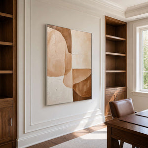 Silver-framed cream and caramel abstract artwork beside wooden bookshelves and a leather chair in a study.