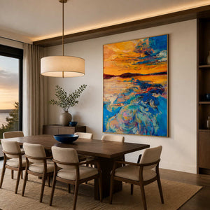Gold framed sunset painting beside a dark dining table overlooking evening water.