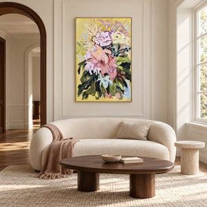 Large pink and cream flowers with dark green leaves on a warm yellow ground in an artwork with a gold-colored frame, displayed in a living room above seating.