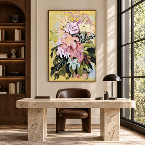 In a study with a desk, an artwork with a brown wood-look frame shows large pink and cream flowers with dark green leaves on a warm yellow ground.