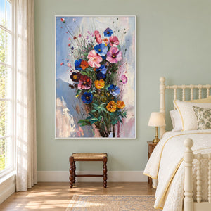 Artwork showing pink, blue and orange flowers on fine stems with dark green leaves against pale blue in a bedroom near the bed, with a white frame.