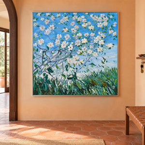 In an entryway above a console or bench, an artwork with a silver-colored frame shows white flowering branches against blue sky above green grass.