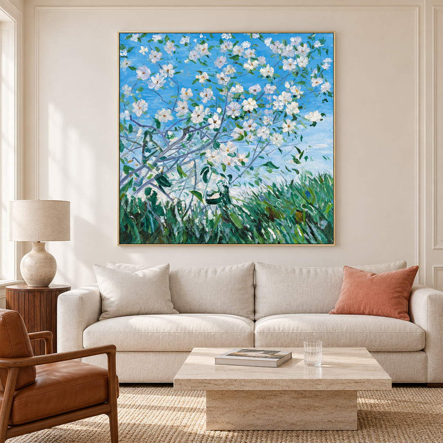 Artwork showing white flowering branches against blue sky above green grass in a living room above seating, with a gold-colored frame.