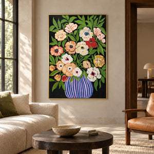 Cream, peach and red flowers with green leaves in a purple-blue striped vase on black in an artwork with a gold-colored frame, displayed in a living room above seating.