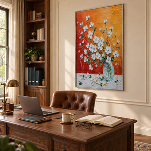 White blossoms in a glass vase against an orange and red background in a silver frame in a home office.