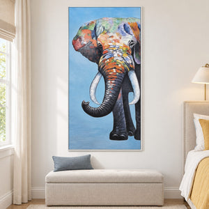 In a bedroom near the bed, an artwork with a white frame shows a front-facing elephant with an upcurled trunk, pale tusks and colored blocks on blue.