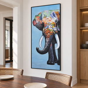 A front-facing elephant with an upcurled trunk, pale tusks and colored blocks on blue in an artwork with a black frame, displayed in a dining area beside the table.