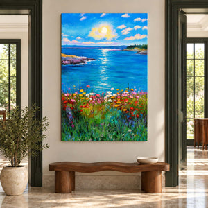 In an entryway above a console or bench, an artwork with no outer frame shows a sunlit blue sea between low shores with colorful flowers in the foreground.