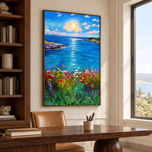 A sunlit blue sea between low shores with colorful flowers in the foreground in an artwork with a brown wood-look frame, displayed in a study with a desk.