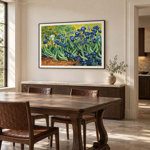 In a dining area beside the table, an artwork with a black frame shows blue flowers and curling green leaves above warm brown soil.