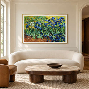 Blue flowers and curling green leaves above warm brown soil in an artwork with a gold-colored frame, displayed in a living room above seating.