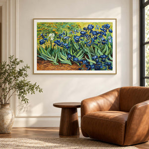 Blue flowers and curling green leaves above warm brown soil in an artwork with a gold-colored frame, displayed beside a reading chair.