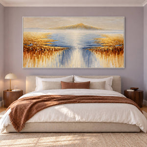 Pale distant mountains above blue-gray water and gold-ochre marks spreading from both sides in an artwork with a white frame, displayed in a bedroom near the bed.