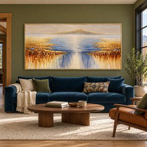 In a living room above seating, an artwork with a gold-colored frame shows pale distant mountains above blue-gray water and gold-ochre marks spreading from both sides.