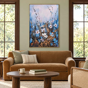 Artwork showing white and blue wildflowers on slender stems beneath a broad pale upper area in a living room above seating, with a gold-colored frame.