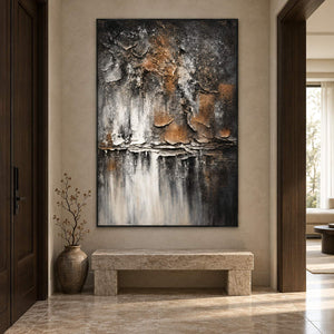 In an entryway above a console or bench, an artwork with a black frame shows dark gray and brown flaking forms above horizontal strata and pale vertical streaks.