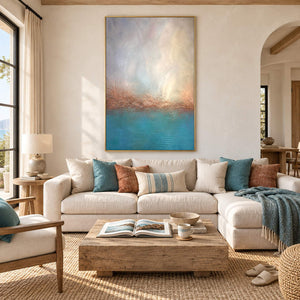 Turquoise coastal painting with a warm shoreline and a luminous pale sky in a gold frame in a living room.