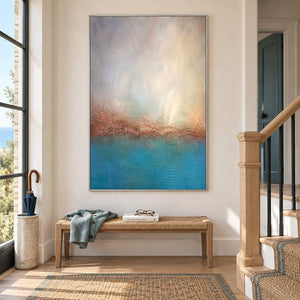 Turquoise coastal painting with a warm shoreline and a luminous pale sky in a silver frame on a stair landing.