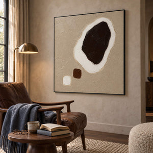 Taupe abstract painting with a large dark brown shape edged in white and two small forms in a black frame in a reading room.