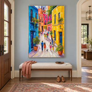 Colorful street painting with people, bright facades, and flowered balconies in a silver frame in an entryway.