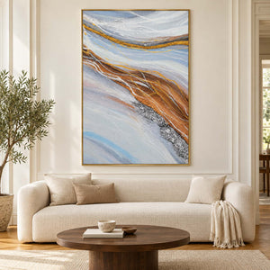 In a living room above seating, an artwork with a gold-colored frame shows pale blue-gray bands curving around ochre areas with fine white lines.