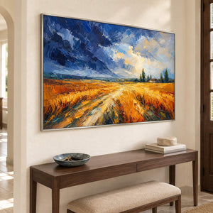 Golden field and winding path painting beneath a blue and cream sky in a silver frame in an entryway.