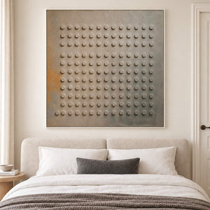 A regular grid of raised round forms on a gray earth-toned ground in a white frame, displayed above a bed.