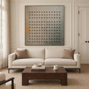 A regular grid of raised round forms on a gray earth-toned ground in a black frame, displayed above a sofa in a living room.