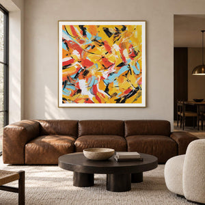 Interlocking yellow, orange, light blue, white and black abstract strokes in an artwork with a gold-colored frame, displayed in a living room above seating.