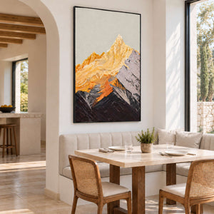 Black-framed artwork of golden peaks and dark mountain ridges above a dining bench, beside a pale table and woven-back chairs.
