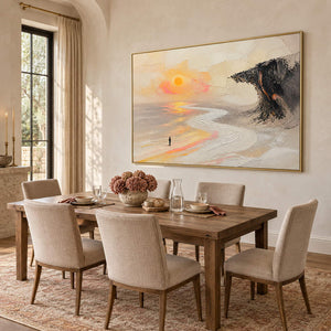 Landscape painting with a lone figure, amber sun, pale shore, and dark cliff in a gold frame in a dining room.