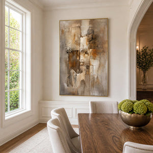 Gold-framed brown and taupe artwork beside a wooden dining table, cream chairs, and bright window.