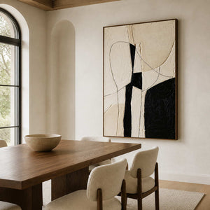 Wood framed ivory and black painting beside a dining table beneath an arched window.