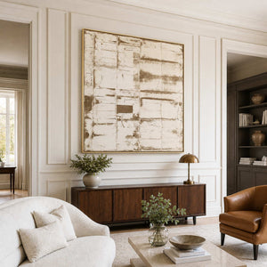 Gold-framed cream abstract above a dark cabinet between ivory and brown living room chairs.