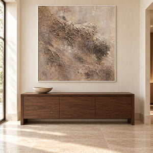 Above a low wooden sideboard, an artwork with a white frame shows irregular beige and taupe relief-like textures on a pale square ground.