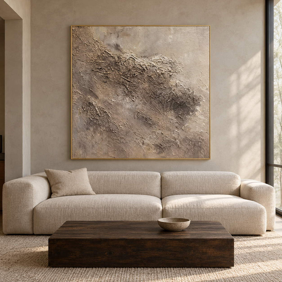 In a living room above seating, an artwork with a gold-colored frame shows irregular beige and taupe relief-like textures on a pale square ground.