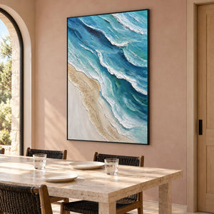 In a dining area beside the table, an artwork with a black frame shows layered blue-green waves with raised white edges beside a pale sandy shore.