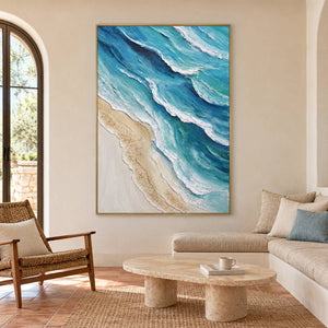Layered blue-green waves with raised white edges beside a pale sandy shore in an artwork with a gold-colored frame, displayed in a living room above seating.