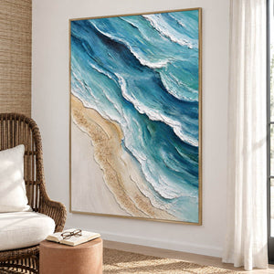 Layered blue-green waves with raised white edges beside a pale sandy shore in an artwork with a gold-colored frame, displayed beside a reading chair.