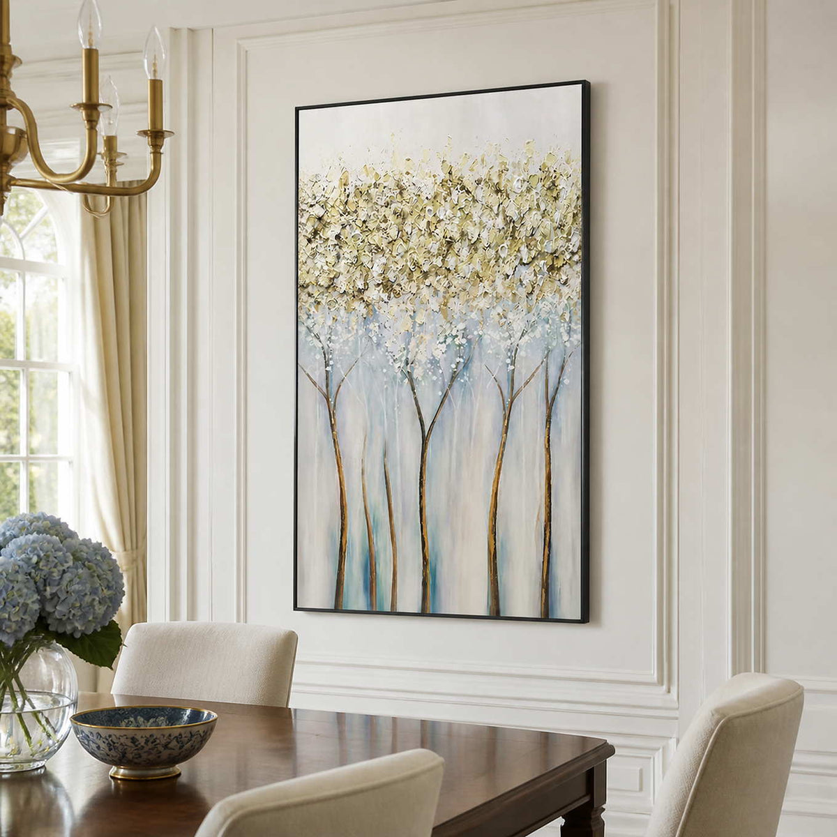 Black-framed delicate tree painting beside a wooden dining table and cream upholstered chairs.