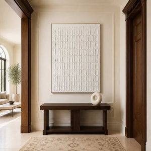 Gold framed cream abstract artwork above a dark wooden console in a bright hallway.