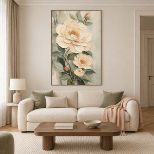 Cream flowers and soft green leaves on a muted background in a brown wood frame, displayed above a sofa in a living room.