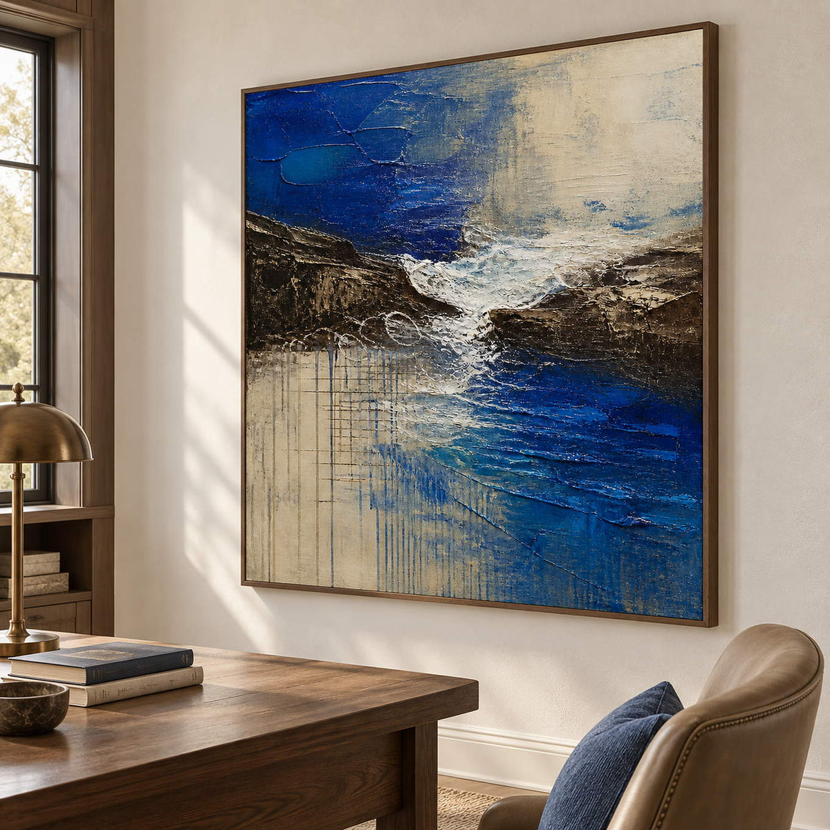 Square cobalt sea, dark rock, and ivory surf textured painting, displayed in a study in a brown wood frame.