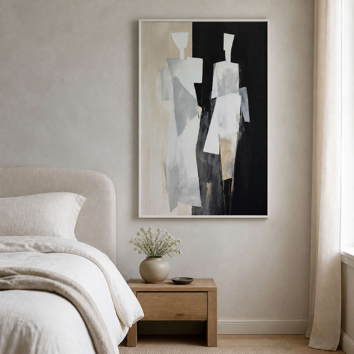 White framed beige and black figurative painting beside a bed and wooden nightstand.