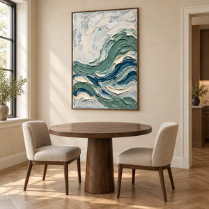 A Sea Breeze in a brown wood frame above a round dining table with light upholstered chairs.
